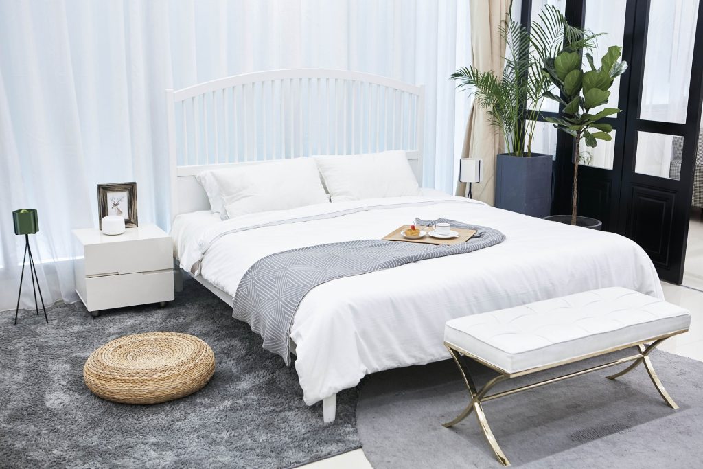 Chambre qui comporte un lit au centre, une verrière, un banc, une table de chevet, un tapis gris et deux grandes plantes